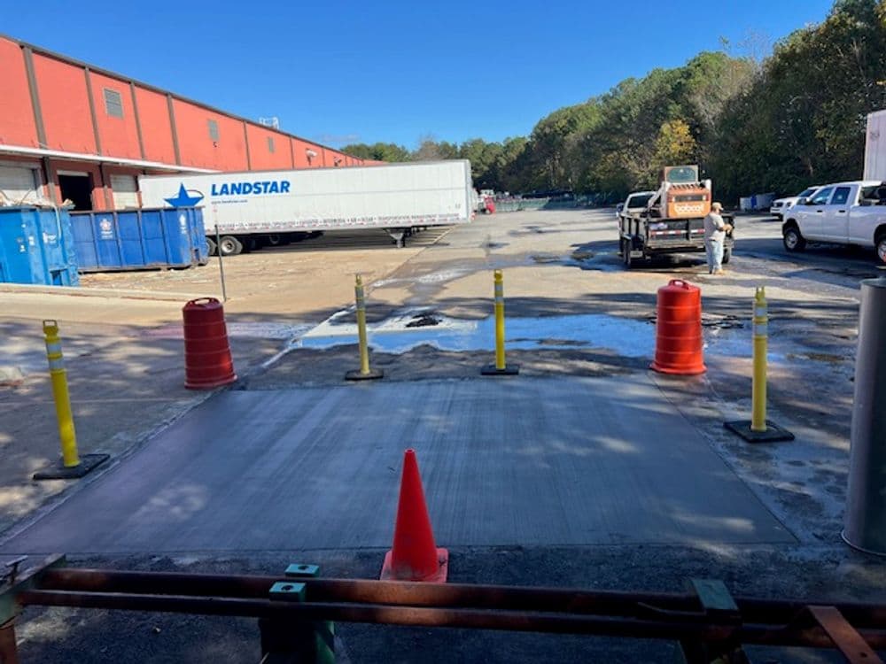 Construction site with freshly poured concrete, safety barriers, and parked trucks in background.