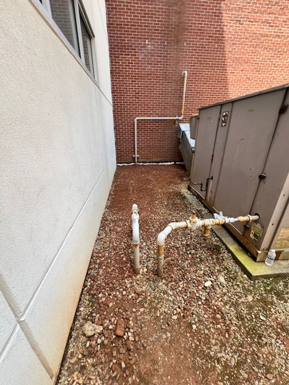 Pipes and equipment beside a building foundation on gravel surface.