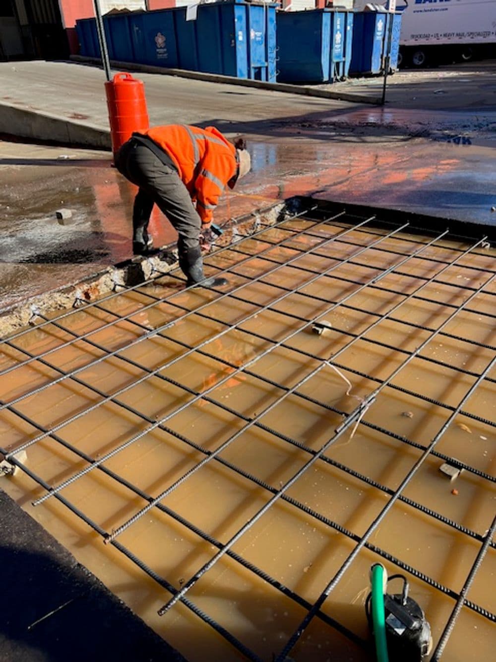 Construction worker installing rebar in a flooded asphalt area with drainage.