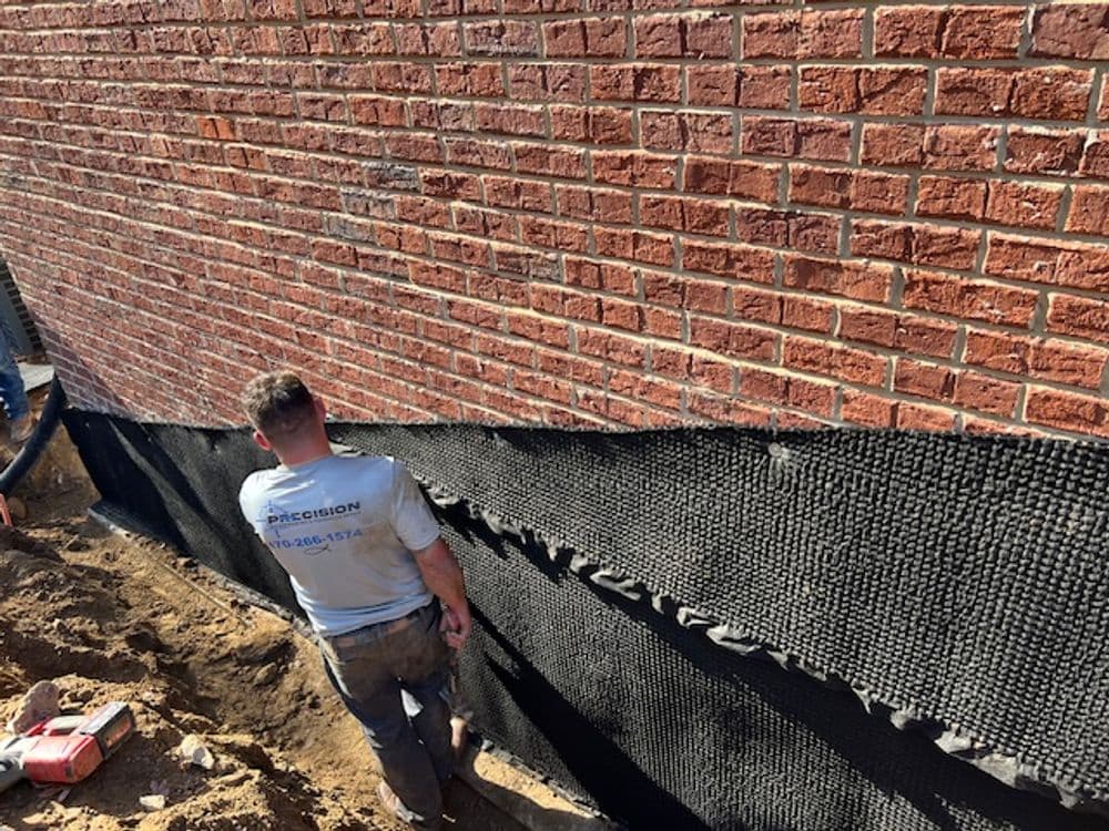 Worker applying foundation drainage system to a brick wall during home construction.