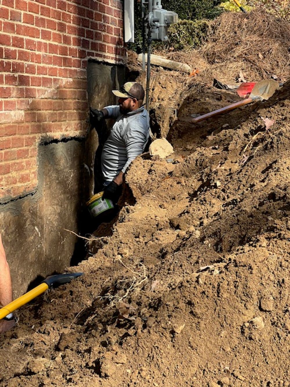 Worker applying waterproof sealant to a brick foundation, surrounded by dug-up soil.