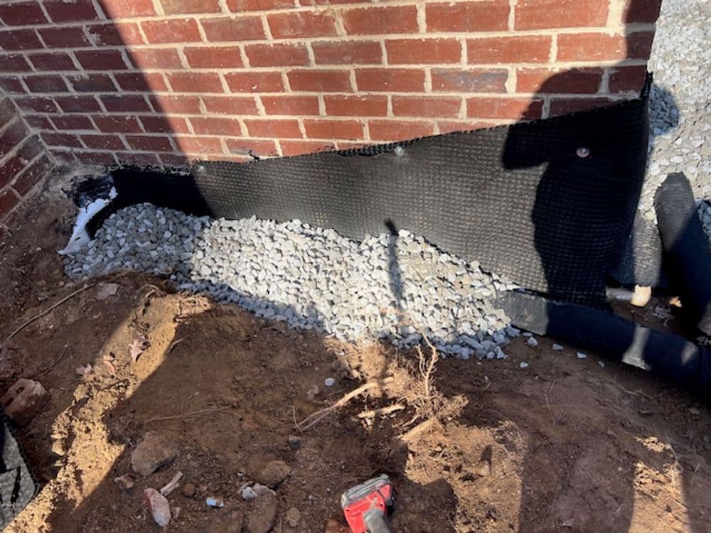 Drainage installation with gravel and landscape fabric against a brick foundation.