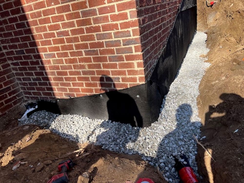 Gravel foundation installation against a brick structure with shadows of workers.