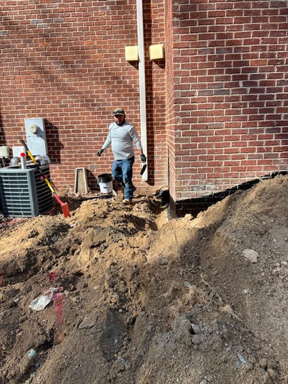 Worker digging near a brick building foundation, with tools and equipment visible.