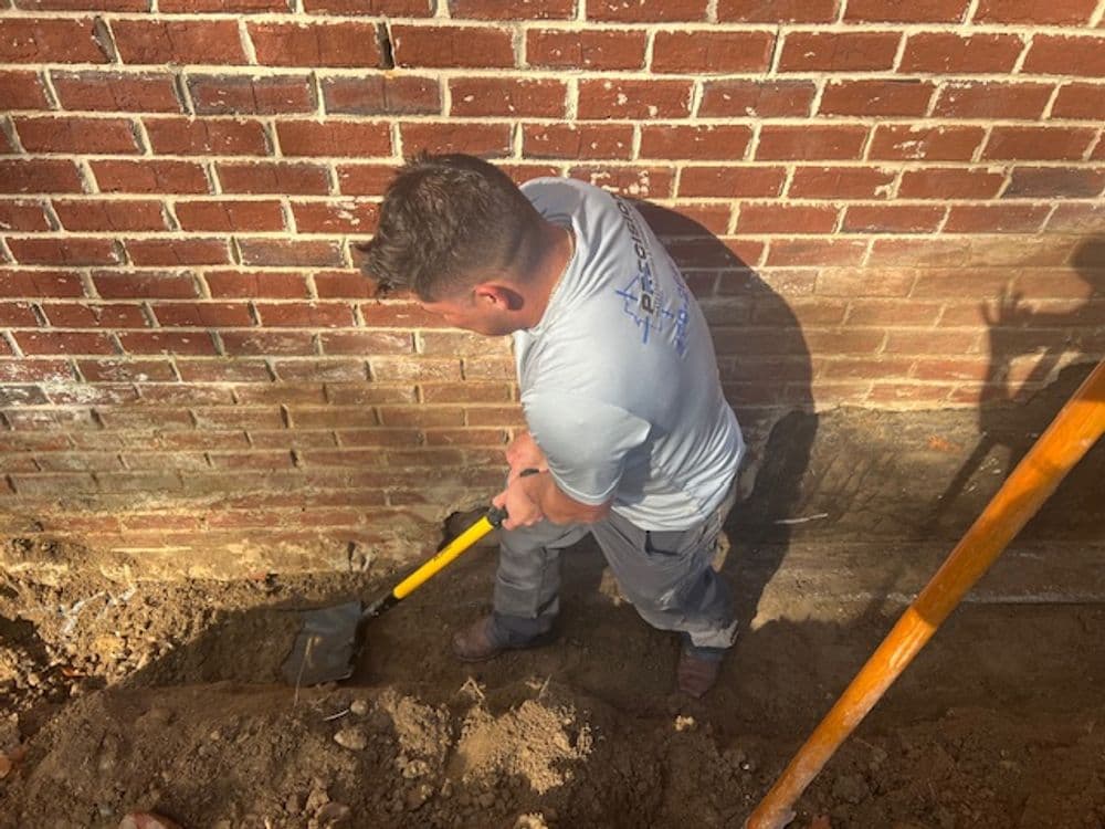 Worker excavating soil near brick wall for construction or repair project.