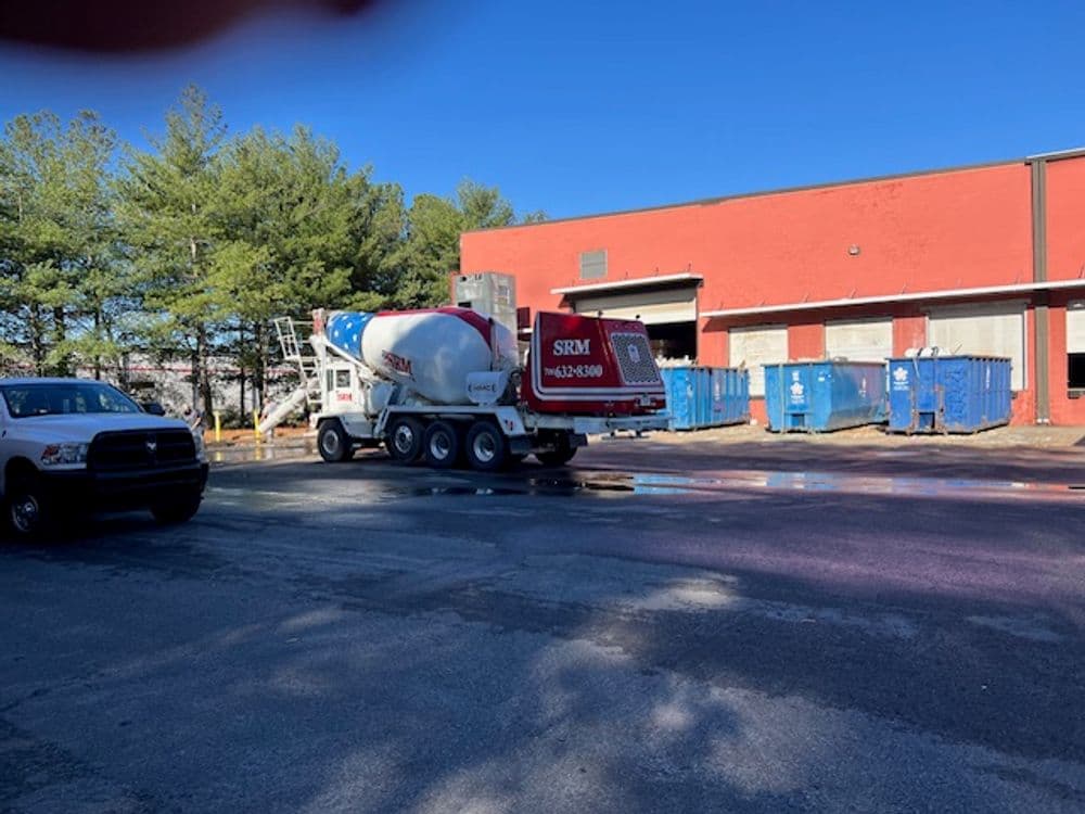 Cement truck parked near a red building and blue dumpsters on a sunny day.