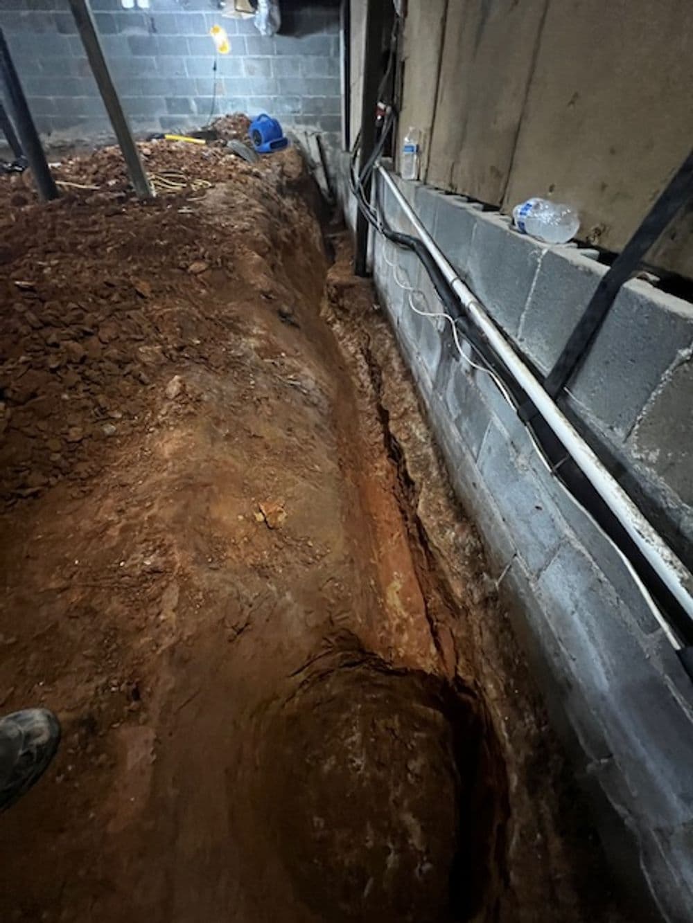 Excavated trench in basement revealing dirt and foundation walls, with visible utilities and debris.