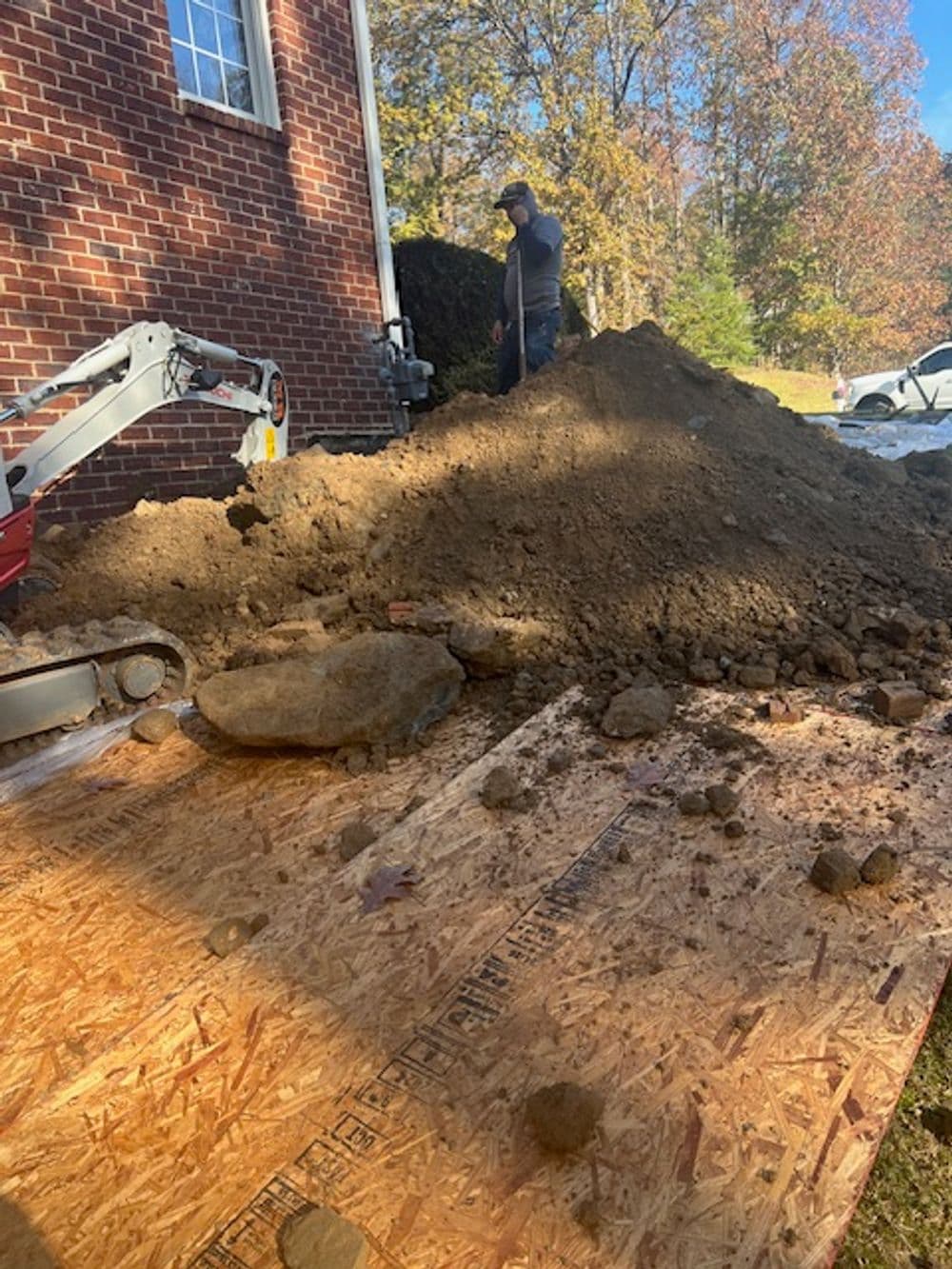Construction worker operating mini excavator on dirt pile near brick house in autumn.