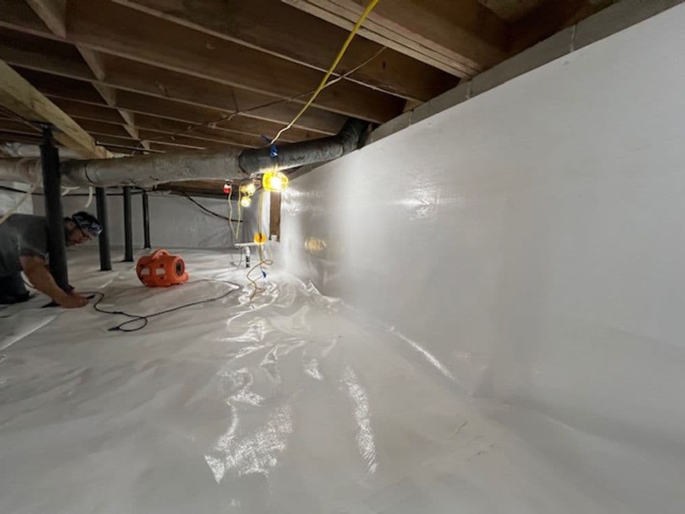 Person inspecting a crawl space with a white vapor barrier and construction lighting.