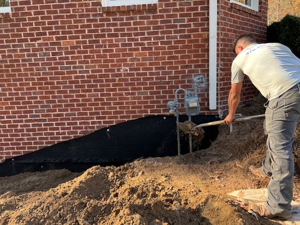 Worker excavating soil near a brick house foundation with gas meters visible.