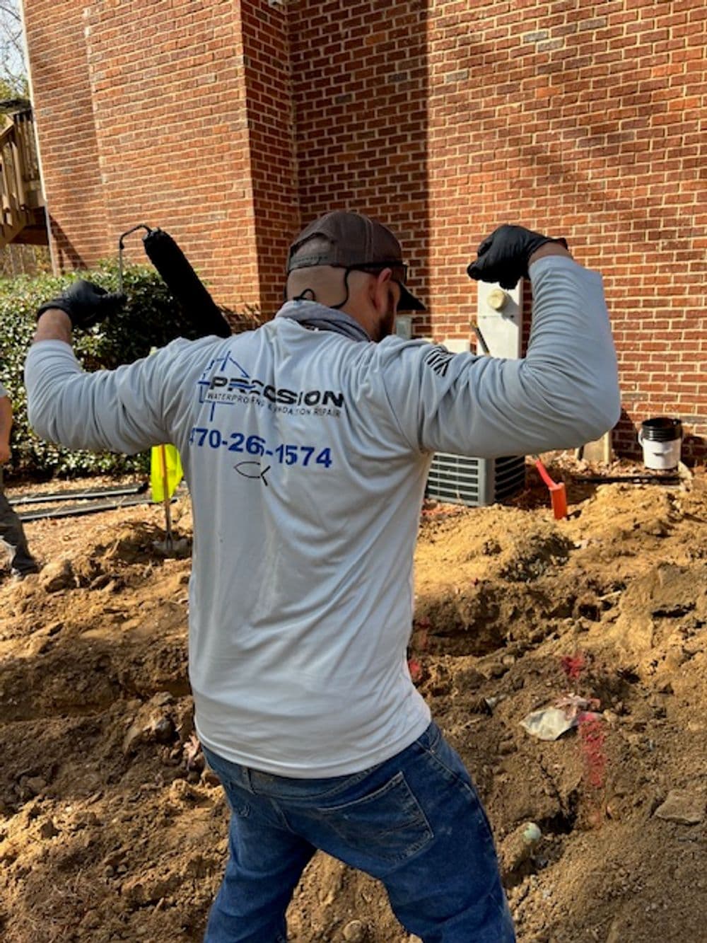 Worker flexing muscles in a gray shirt with a business logo and phone number on a construction site.