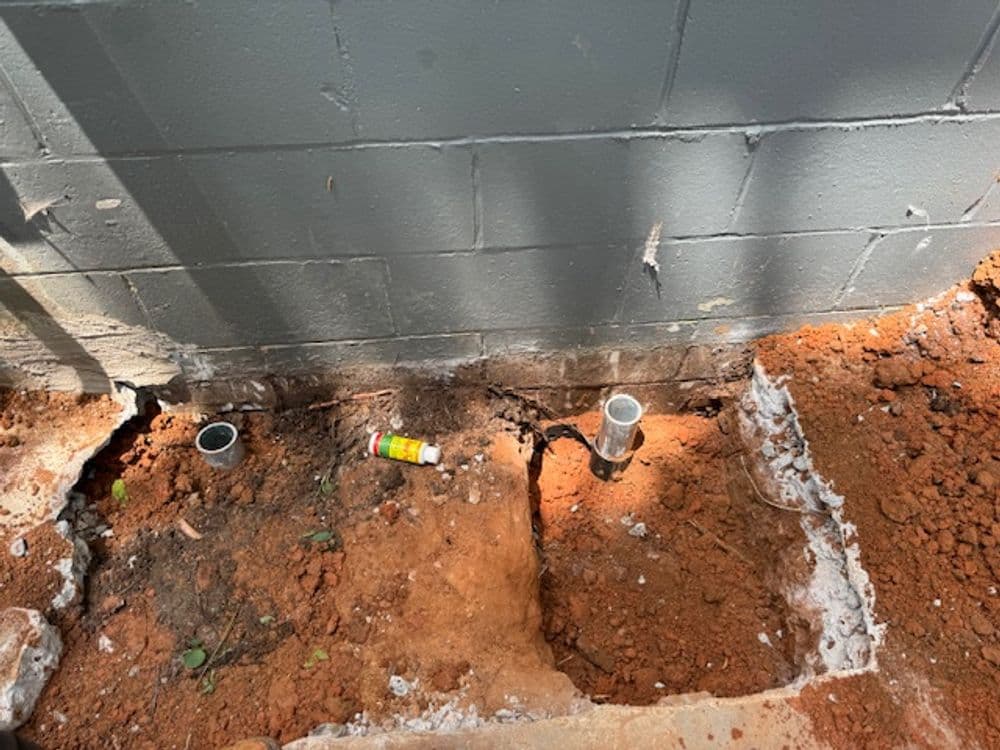 Excavated area with exposed pipes and soil near a gray wall, indicating plumbing work.