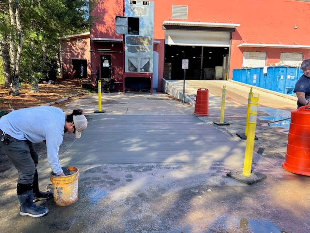 Worker applying concrete in a commercial area with safety barriers and a loading dock in background.