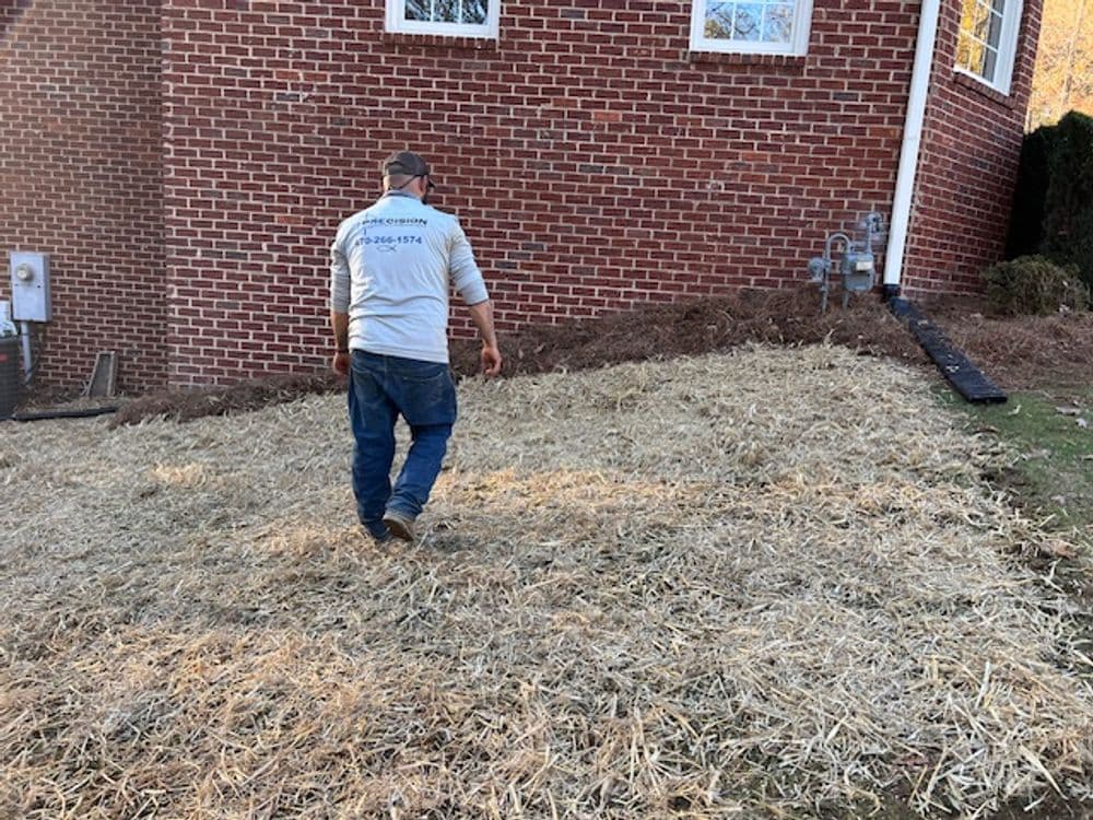 Worker spreading straw mulch on freshly graded soil near a brick home exterior.