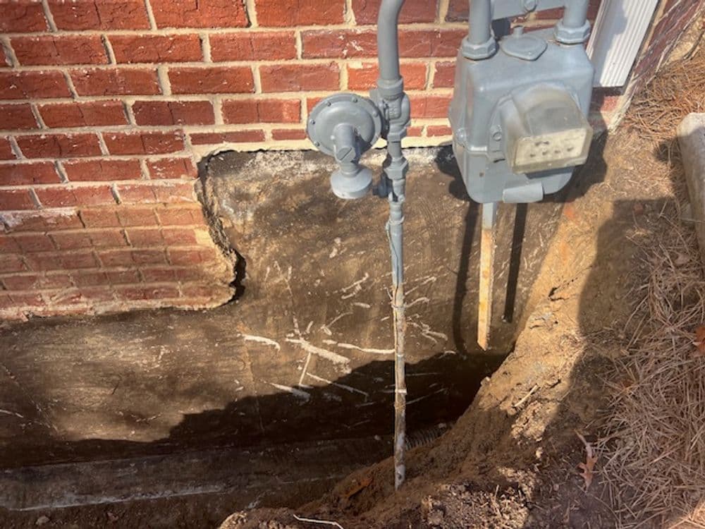 Gas meter installation next to a brick foundation with exposed earth and utility lines.