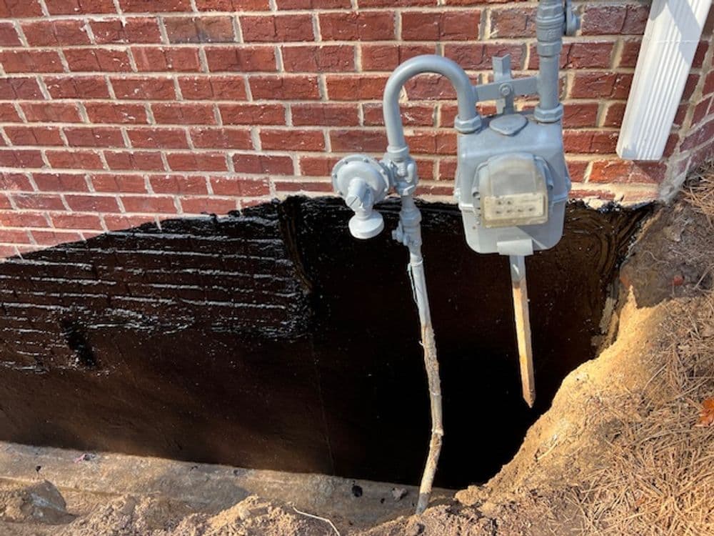 Gas meter mounted on a brick wall with exposed trench and waterproofing membrane.
