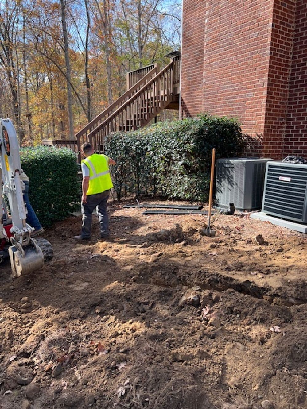 Construction worker in yellow vest digging near a brick building and air conditioning unit.