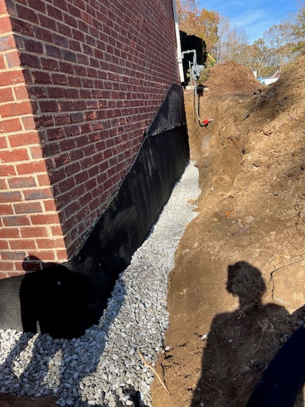Excavated trench beside brick house filled with gravel for drainage system installation.