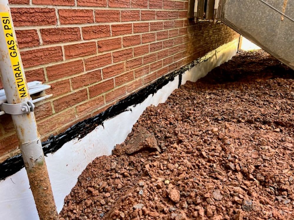 Exposed foundation with waterproofing membrane and gravel for drainage near brick wall.