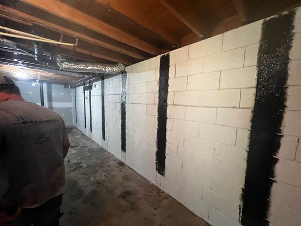 Man applying waterproof sealant to basement walls with unfinished ceiling and concrete floor.