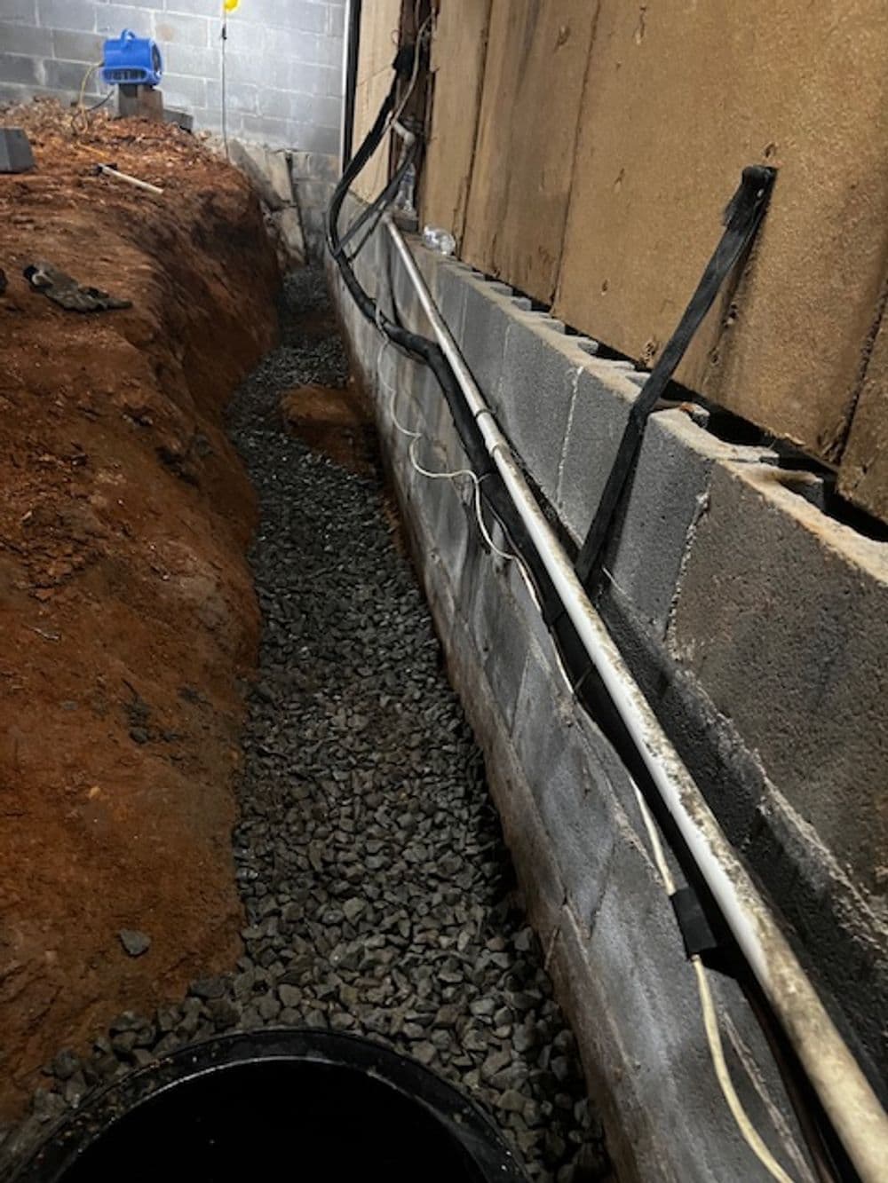 Gravel trench alongside a foundation wall with exposed drainage pipes and electrical lines.