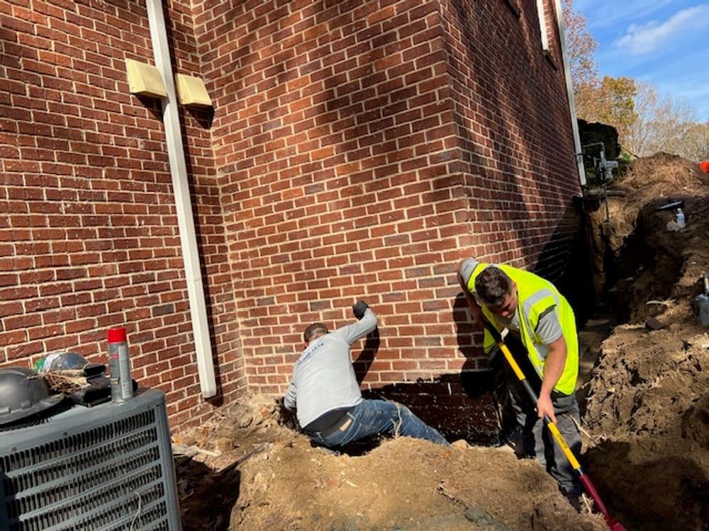 Two workers excavate soil near a brick building for construction repairs.