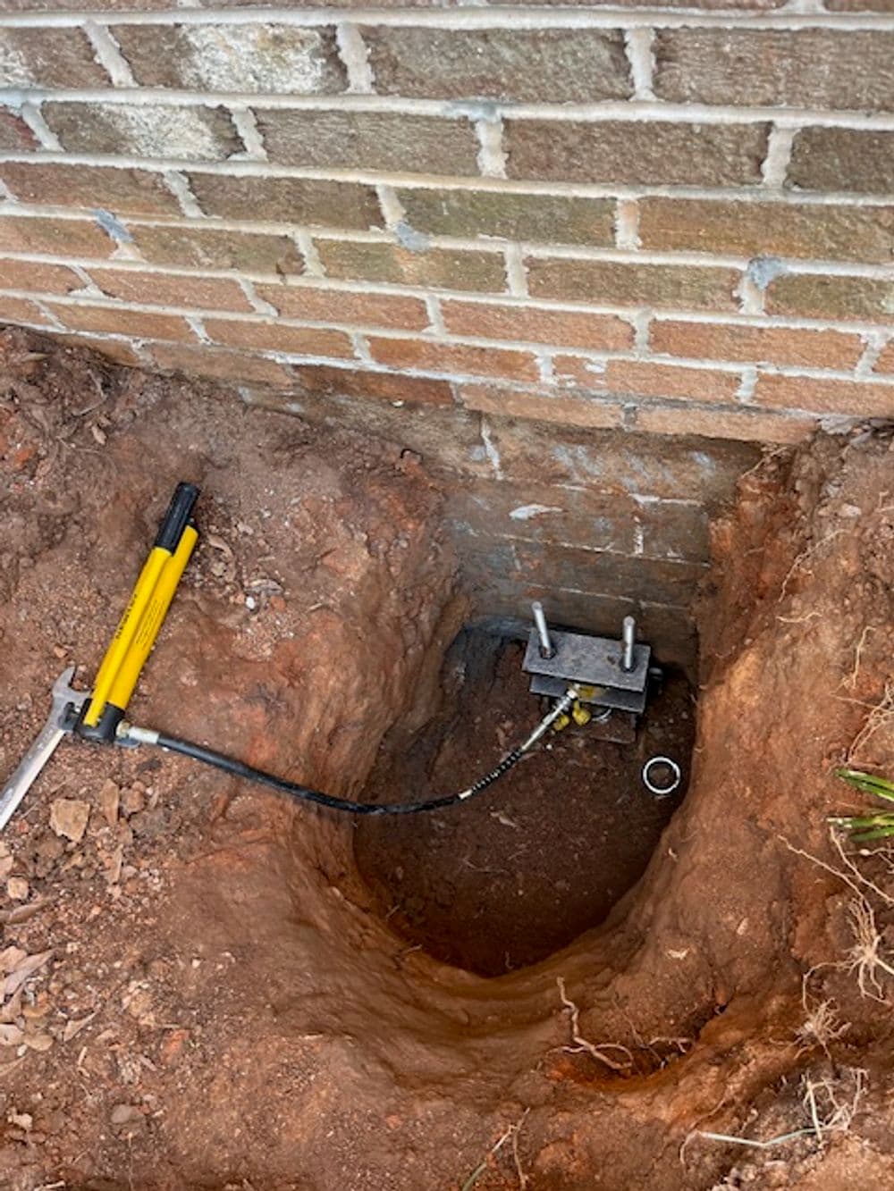 Foundation repair with hydraulic lifting equipment in a dug hole near a brick wall.