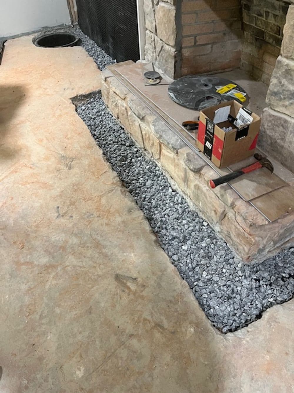 Renovation scene showing gravel base and stone fireplace with tools and a box nearby.